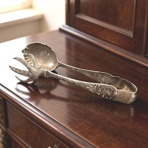 May include: A pair of silver-toned metal serving tongs featuring a decorative handle with intricate floral engravings and a unique claw-shaped end on one side, paired with a scalloped, perforated bowl-shaped end on the opposite side.