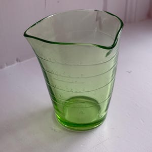 May include: A vintage, light green glass measuring cup with a flared top and a pouring spout. The cup has etched measurement markings in cups and fluid ounces. The glass is transparent, allowing the contents to be easily seen.
