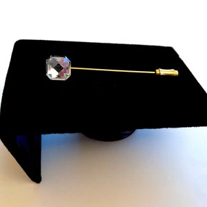 May include: A gold tie tack with a clear, faceted, square-shaped crystal.
