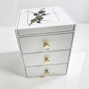 May include: A white storage chest with three drawers, each featuring a round gold-toned knob. The top surface has a rectangular panel with a black and gold floral illustration of iris flowers against a white background with fine vertical lines.
