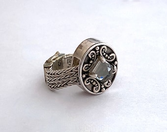 May include: A silver-toned ring with an adjustable band and a decorative, circular face. The face features a square, gray gemstone surrounded by ornate scrollwork. The band has a woven design, adding texture to the jewelry.