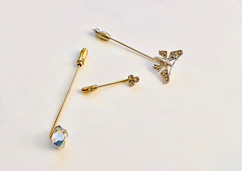 May include: Three decorative pins in gold and silver. One pin features a clear crystal, one has a cluster of small crystals, and the third is a silver butterfly with crystal accents.