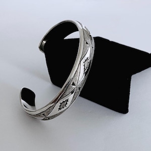 May include: A silver-toned cuff bracelet featuring a polished finish with a repeating pattern of etched diamonds and triangles. The metal surface displays a contrast between smooth, reflective areas and textured, matte-finished geometric shapes.