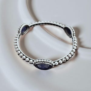 May include: A silver bangle bracelet featuring a beaded texture along the band and three oval-shaped, dark blue faceted stones set at intervals. The metal has a polished finish with darkened detailing between the beads to highlight the texture.