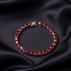 May include: A tennis bracelet featuring a continuous row of oval-cut, deep red gemstones set in a metallic frame. The bracelet is secured with a rectangular box clasp mechanism, showcasing a uniform arrangement of vibrant red stones in a circular design.