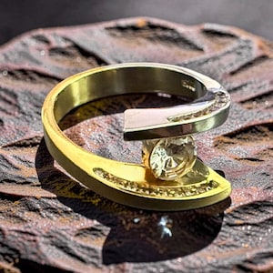 May include: A bypass ring featuring a two-tone design with a polished gold-colored band and a silver-colored upper section. A single round, clear faceted stone is set between the two ends of the band, which are accented with small embedded stones.