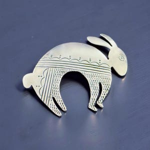 May include: A silver-coloured metal brooch shaped like a rabbit. The rabbit features etched details, including lines and dots, and is posed in a curved position. The brooch is likely a decorative accessory.
