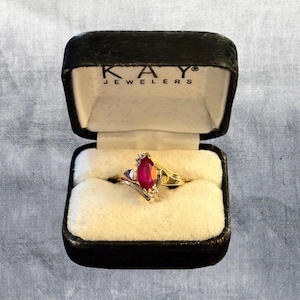 May include: A gold ring featuring a marquise-cut red gemstone centered between small clear stones, presented in a black Kay Jewelers box with the brand name printed in white on the interior lid lining.