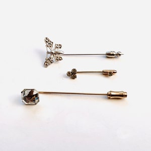 May include: Three silver and gold stick pins with decorative tops. One pin features a butterfly design with clear rhinestones. Another pin features a cluster of clear rhinestones. The third pin features a large, clear, square-shaped rhinestone.