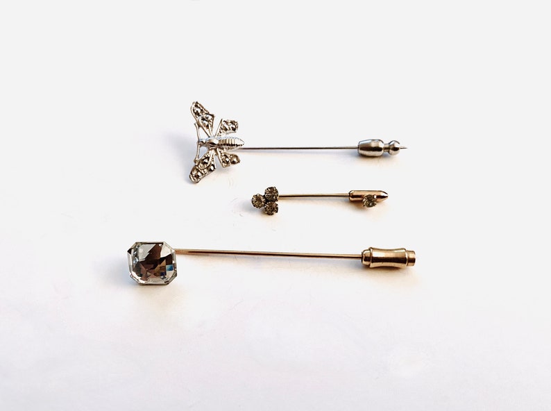 May include: Three silver and gold stick pins with decorative tops. One pin features a butterfly design with clear rhinestones. Another pin features a cluster of clear rhinestones. The third pin features a large, clear, square-shaped rhinestone.