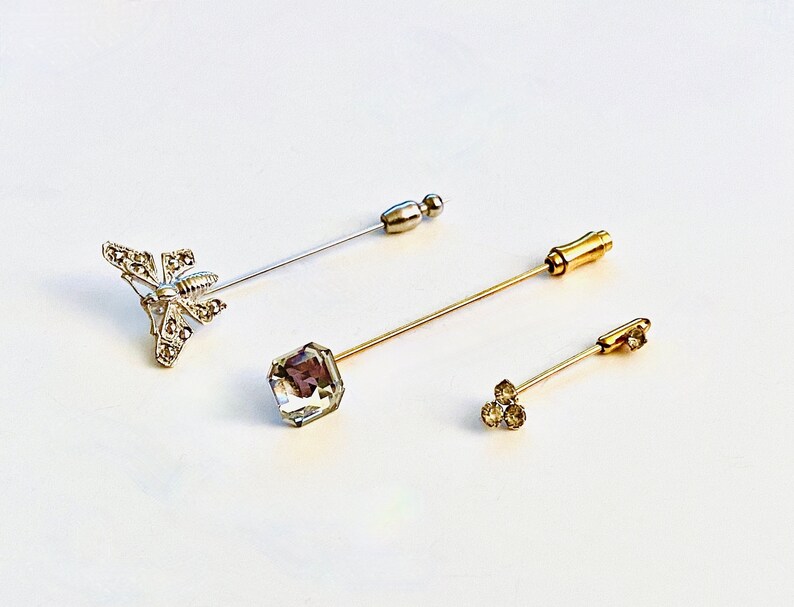 May include: Three decorative pins with different embellishments. The first pin has a silver butterfly design, the second pin has a clear square crystal, and the third pin has three small clear crystals.