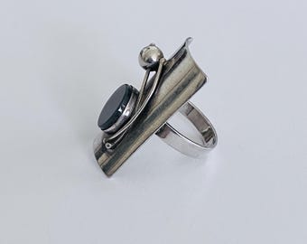 May include: A silver ring with a black onyx stone and a silver orb. The ring has a unique, abstract design.