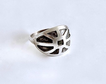 May include: A silver-toned ring featuring a wide, rounded face with a cut-out geometric pattern. The design consists of a central square surrounded by radiating segments, creating a series of open spaces that reveal a dark, recessed background.