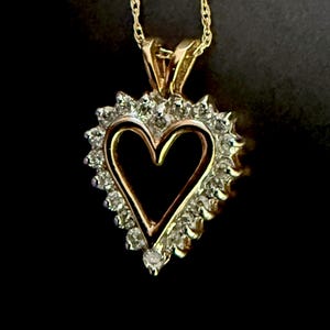 May include: A gold heart-shaped pendant necklace featuring a central open heart design surrounded by a border of small, clear, round-cut stones. The pendant is suspended from a delicate gold rope chain, creating a classic and elegant jewelry piece.