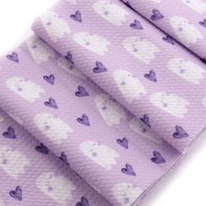 May include: A folded fabric with a repeating pattern of white ghosts and purple hearts on a light purple background. The ghosts have closed eyes and small blush marks. The fabric is textured.