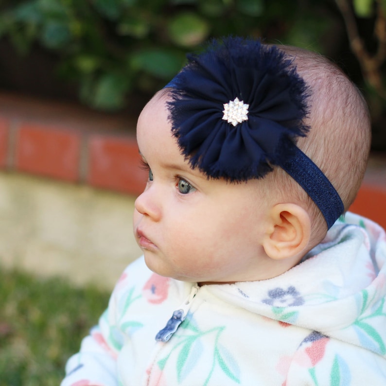 May include: A baby wearing a white floral print hooded jacket with a navy blue flower headband with a rhinestone center.