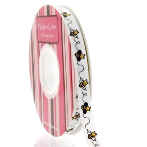 May include: A spool of white ribbon with a repeating pattern of black and yellow bees. The ribbon is packaged on a pink and brown striped spool with the text "Hair Bow Center" and "The happy place".