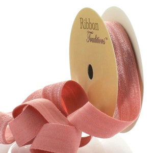 May include: A spool of dusty rose pink satin ribbon with a shimmer finish. The ribbon is wrapped around a white and beige spool with the text "Ribbon Traditions" printed on it.