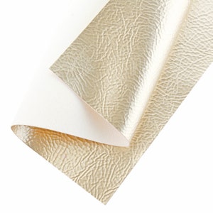 May include: A close-up of a gold faux leather fabric with a textured surface. The fabric is folded in half, revealing a white backing.
