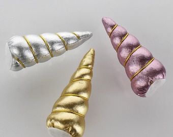 May include: Three unicorn horn headbands in gold, silver, and pink. Each headband is made of fabric with a spiral design and gold trim.