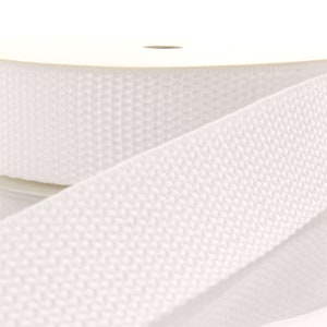 May include: White hook and loop tape, also known as Velcro, is shown rolled up on a spool. The tape has a textured surface and is designed for attaching two pieces of fabric together.