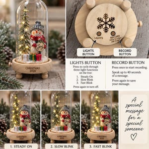 Pode incluir: Uma cúpula de vidro contendo um boneco de neve, uma árvore de Natal em miniatura com luzes e pequenos presentes sobre uma base de madeira com a etiqueta CHRISTMAS 2026. A base possui um ícone de floco de neve, botão de luz e gravação.