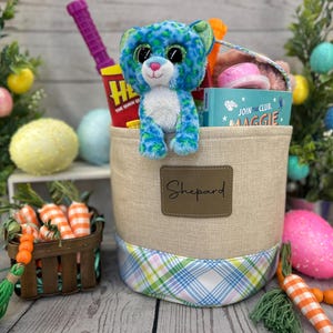 May include: A beige canvas Easter basket with a blue and green plaid trim. The basket is filled with a blue and green plush toy, a book titled "Join the Club, Maggie", a pink toy, and a purple toy. The basket has a brown leather tag with the name "Sheppard" written on it.