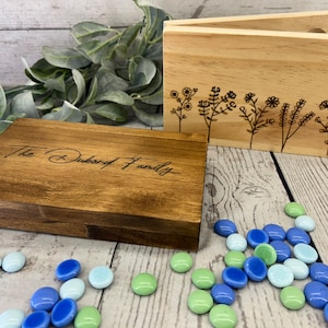 Peut inclure: Deux boîtes en bois avec des motifs gravés. La plus petite boîte a un motif floral et la plus grande boîte porte le texte "The Dubord Family". Des cailloux de verre bleus et verts sont éparpillés devant les boîtes.