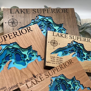 Puede incluir: Tres placas de madera con un mapa en relieve del Lago Superior. Las placas est&aacute;n hechas de diferentes tipos de madera y tienen una rosa de los vientos y texto con las dimensiones del lago. El texto dice: "Lago Superior", "Longitud: 348 millas, Ancho: 160 millas, Profundidad: 1332 pies, &Aacute;rea de superficie: 31,700" y "17.04996&deg; N, -86.50245&deg; E".