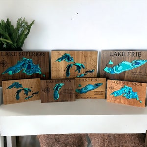 Puede incluir: Seis placas de madera con mapas de los Grandes Lagos cortados con l&aacute;ser. Cada placa presenta un lago diferente: Superior, Michigan, Erie y Ontario. Las placas est&aacute;n te&ntilde;idas de un color marr&oacute;n claro y tienen un mapa azul verdoso del lago con curvas de nivel. Las placas est&aacute;n dispuestas en fila en un estante blanco.