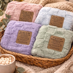 May include: Four plush blankets in a woven basket, each a different pastel color: pink, blue, purple, and green. Each blanket has a rectangular patch with text. A cup of hot chocolate and cookies are in the foreground.