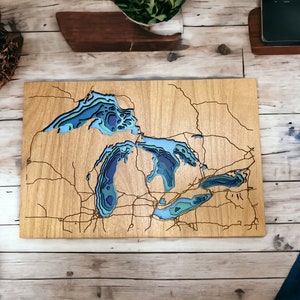 Puede incluir: Una pieza de arte de pared de madera que representa los Grandes Lagos en un estilo 3D en capas. Los lagos est&aacute;n tallados en la madera y pintados en tonos de azul y verde. La tierra circundante se muestra en un tono de madera marr&oacute;n claro.