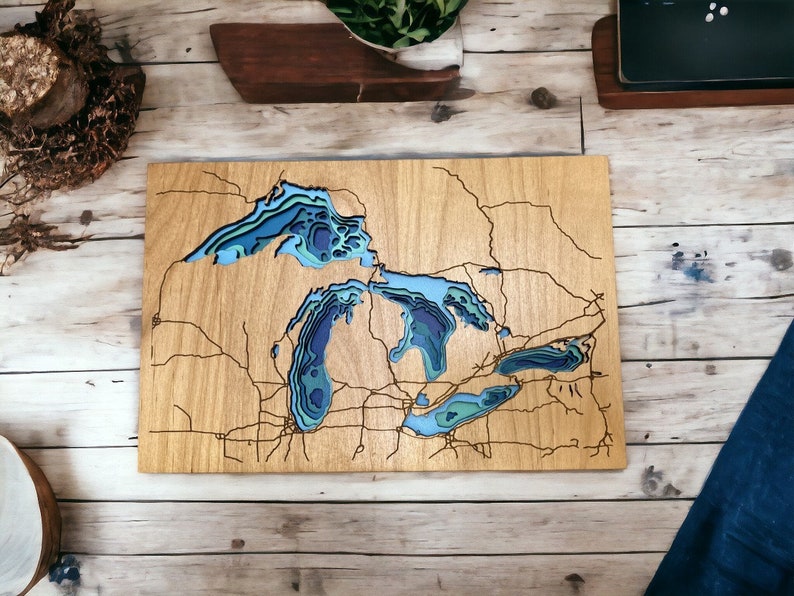 Puede incluir: Una pieza de arte de pared de madera que representa los Grandes Lagos en un estilo 3D en capas. Los lagos est&aacute;n tallados en la madera y pintados en tonos de azul y verde. La tierra circundante se muestra en un tono de madera marr&oacute;n claro.
