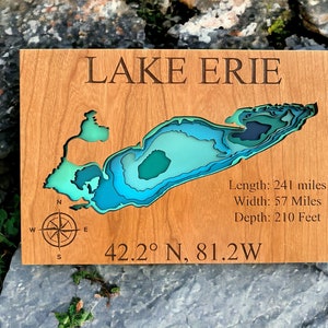 Puede incluir: Un mapa de madera del lago Erie con una rosa de los vientos y las coordenadas 42.2&deg; N, 81.2&deg; O. El mapa est&aacute; en capas de colores azul, verde y turquesa para representar la profundidad del lago. El texto "LAKE ERIE" est&aacute; en la parte superior del mapa. El texto "Longitud: 241 millas, Ancho: 57 millas, Profundidad: 210 pies" est&aacute; en el lado derecho del mapa.