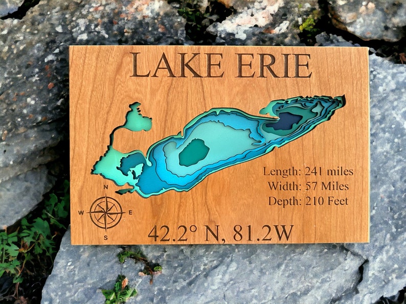 Puede incluir: Un mapa de madera del lago Erie con una rosa de los vientos y las coordenadas 42.2&deg; N, 81.2&deg; O. El mapa est&aacute; en capas de colores azul, verde y turquesa para representar la profundidad del lago. El texto "LAKE ERIE" est&aacute; en la parte superior del mapa. El texto "Longitud: 241 millas, Ancho: 57 millas, Profundidad: 210 pies" est&aacute; en el lado derecho del mapa.