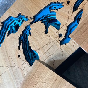 Puede incluir: Una pieza de arte de pared de madera que representa los Grandes Lagos en un estilo en capas 3D. Los lagos est&aacute;n pintados en tonos de azul y negro, con la tierra circundante en un tono de madera natural.