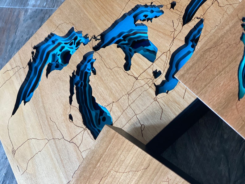 Puede incluir: Una pieza de arte de pared de madera que representa los Grandes Lagos en un estilo en capas 3D. Los lagos est&aacute;n pintados en tonos de azul y negro, con la tierra circundante en un tono de madera natural.