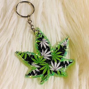 May include: A green and white marijuana leaf keychain with glitter accents. The keychain is made of resin and has a silver metal ring.