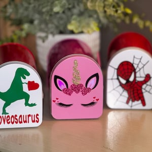 Puede incluir: Tres pequeñas cajas de metal de colores con diferentes diseños. La caja de la izquierda tiene un dinosaurio verde que sostiene un corazón rojo y el texto "Lovesaurus". La caja del medio tiene un unicornio rosa con purpurina y corazones rosas. La caja de la derecha tiene un personaje de Spiderman rojo con un diseño de telaraña.