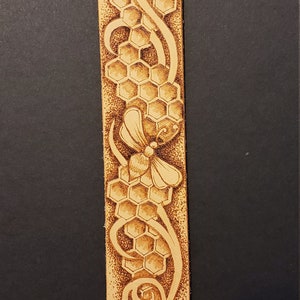 May include: A light brown leather bracelet with a detailed design of a bee and honeycomb. The bracelet has a silver snap closure.