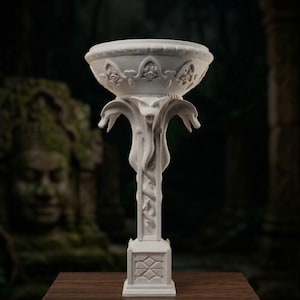 May include: A white, ornate pedestal bowl with a wide, shallow basin. The bowl is supported by a detailed column with stylized floral and serpent motifs. The base is square, and the entire piece is set on a wooden surface.