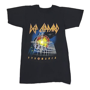 Könnte beinhalten: Schwarzes T-Shirt mit einem Aufdruck des Albumcovers von Def Leppards Pyromania. Der Aufdruck zeigt eine Stadtlandschaft mit einem großen, leuchtenden Kreis in der Mitte.