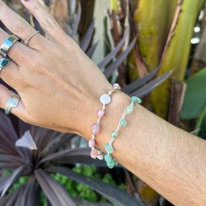 以下が含まれることがあります： ピンク、緑、白の石を使った2つのビーズブレスレットが手首に着用されています。ブレスレットは白いコードに通されています。また、銀色の指輪もいくつか着用しています。背景には緑と紫の植物があります。