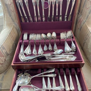 May include: A wooden silverware chest with a deep purple velvet lining, filled with a complete set of silver-colored flatware. The set includes forks, knives, spoons, and serving utensils, all neatly arranged in the open drawers.