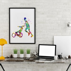 May include: A watercolor painting of a person pushing a child in a stroller. The painting is in a black frame and is hanging on a white brick wall. There is a laptop, a lamp, and a cup on a wooden table in front of the painting.