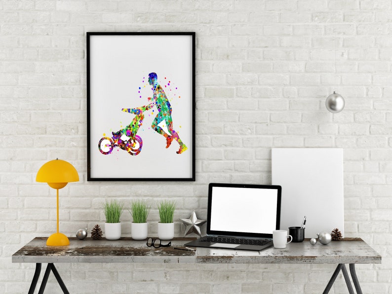 May include: A watercolor painting of a person pushing a child in a stroller. The painting is in a black frame and is hanging on a white brick wall. There is a laptop, a lamp, and a cup on a wooden table in front of the painting.