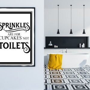 May include: Black and white framed bathroom sign with the text "Sprinkles are for cupcakes not toilets". The sign is hanging on a white wall in a bathroom with a white bathtub, yellow towel, and black and white tiled floor.