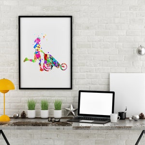 May include: A colorful watercolor painting of a person pushing a wheelbarrow. The painting is framed in black and hanging on a white brick wall. A laptop, a lamp, and plants are on a wooden table in front of the painting.