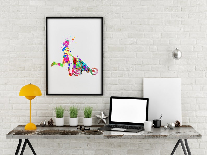 May include: A colorful watercolor painting of a person pushing a wheelbarrow. The painting is framed in black and hanging on a white brick wall. A laptop, a lamp, and plants are on a wooden table in front of the painting.