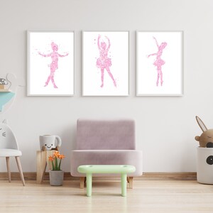 May include: Three framed prints of pink ballerina silhouettes on a white background. The prints are hanging on a white wall in a child's room. The room also features a light green stool, a white cat-shaped chair, and a gray storage bin with a panda face.